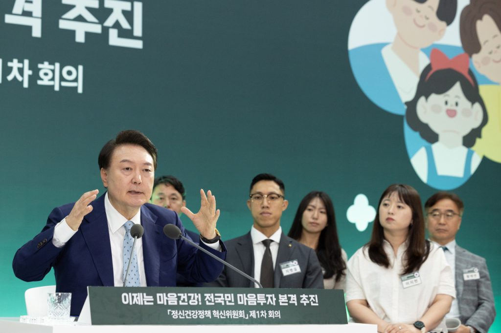 Korean man raises hands in emphasis while speaking to panel of people.